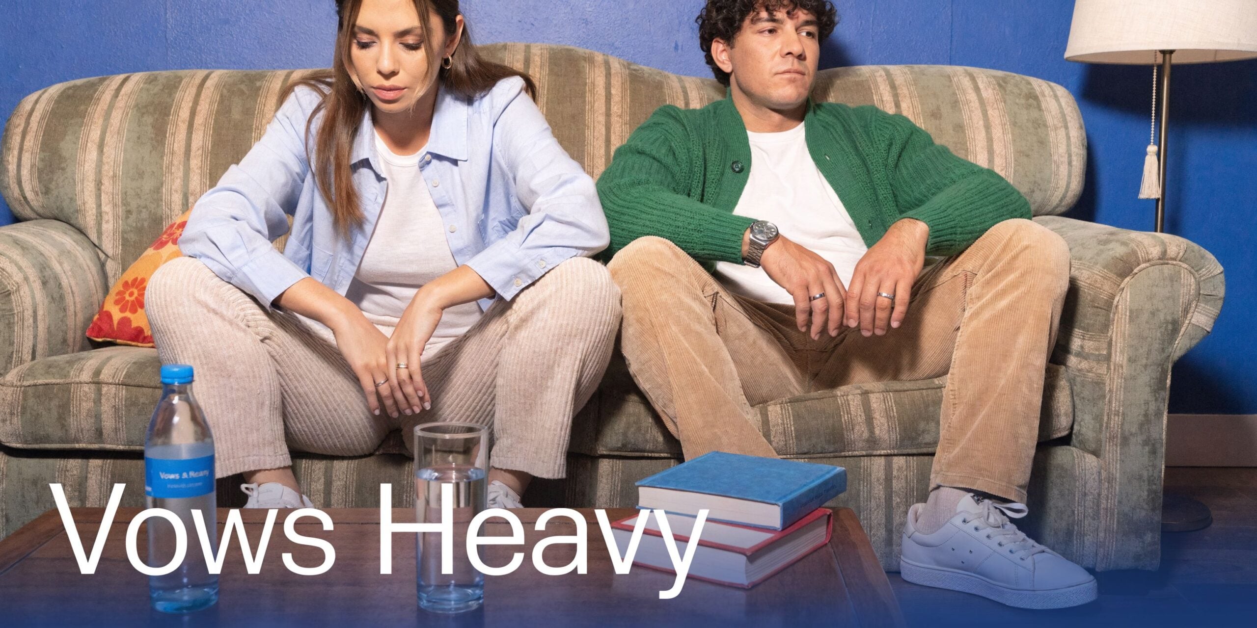 A couple sits apart on a sofa in a dimly lit room, appearing disconnected and somber.