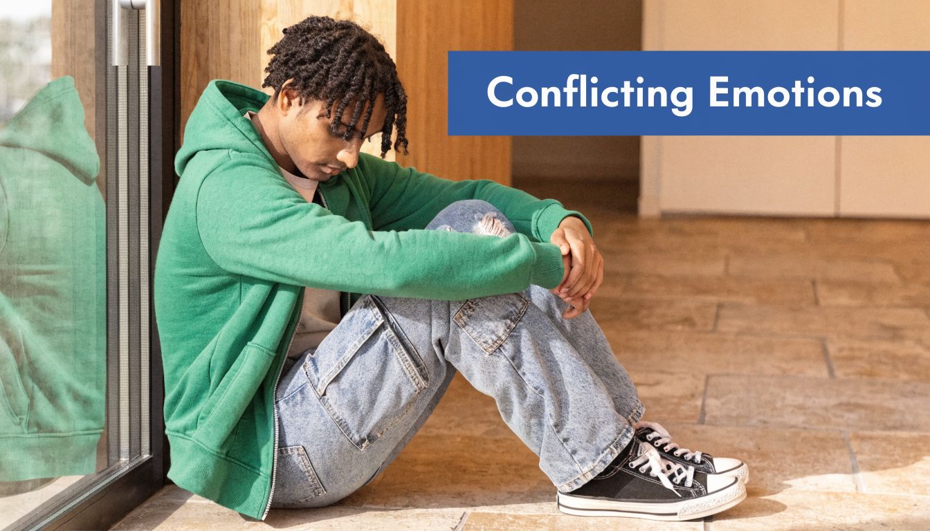 A young person with dreadlocks sitting on the floor in a green hoodie, appearing pensive and sad.