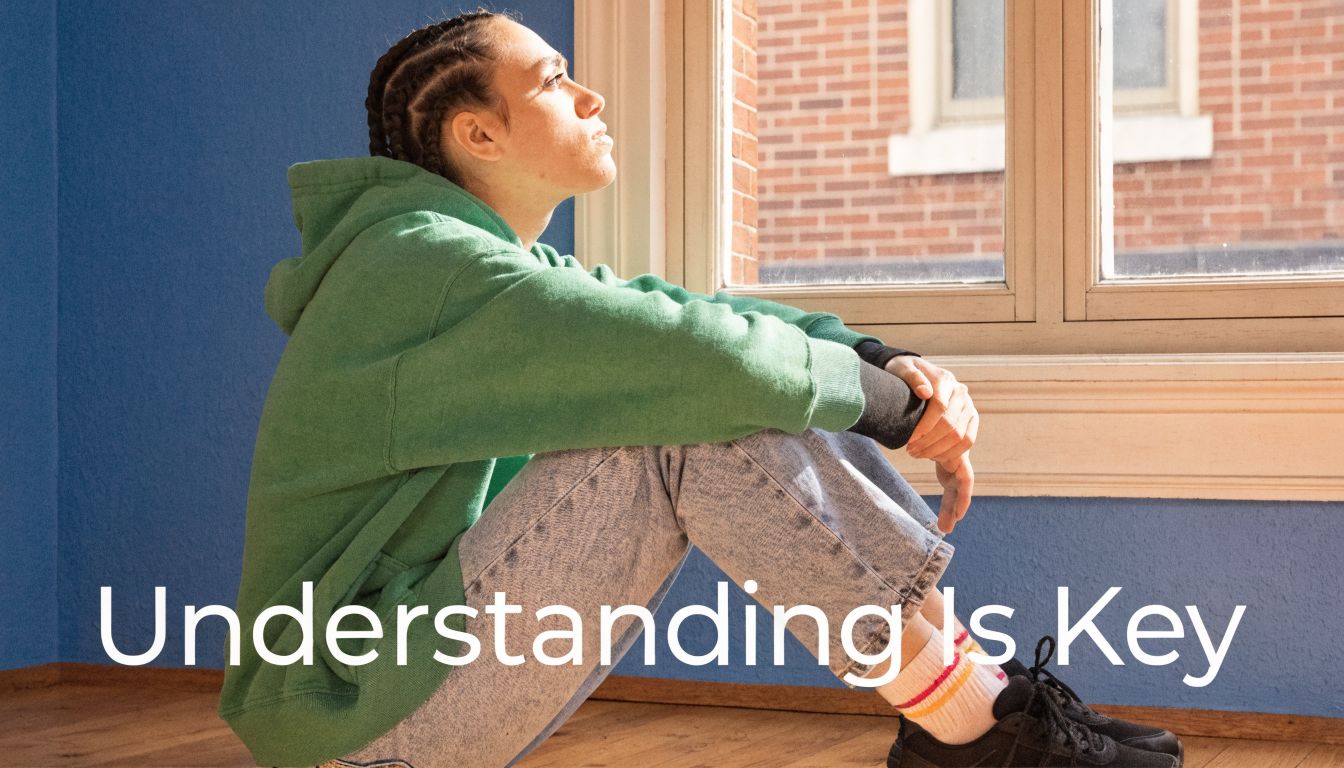 A pensive young person with braided hair sitting on the floor by a window looking away.